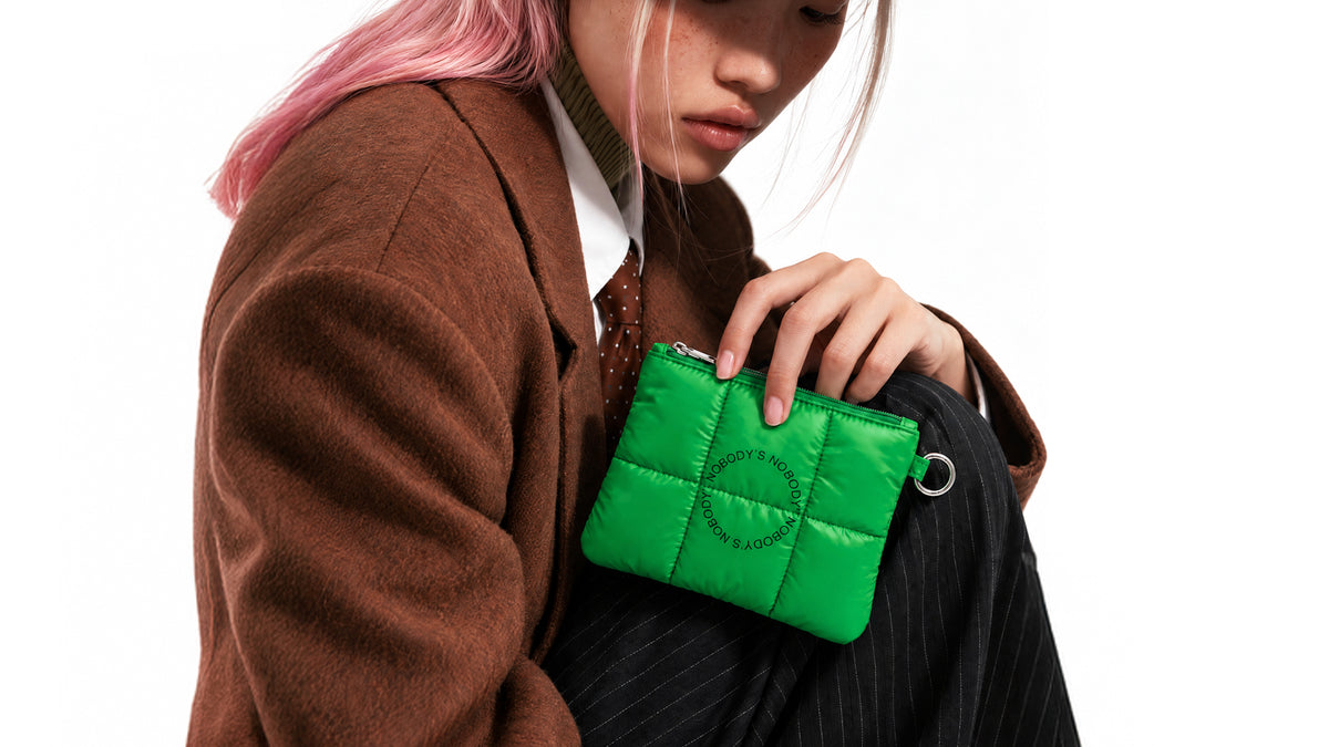 Model in a brown coat holding the green quilted NBDY travel pouch